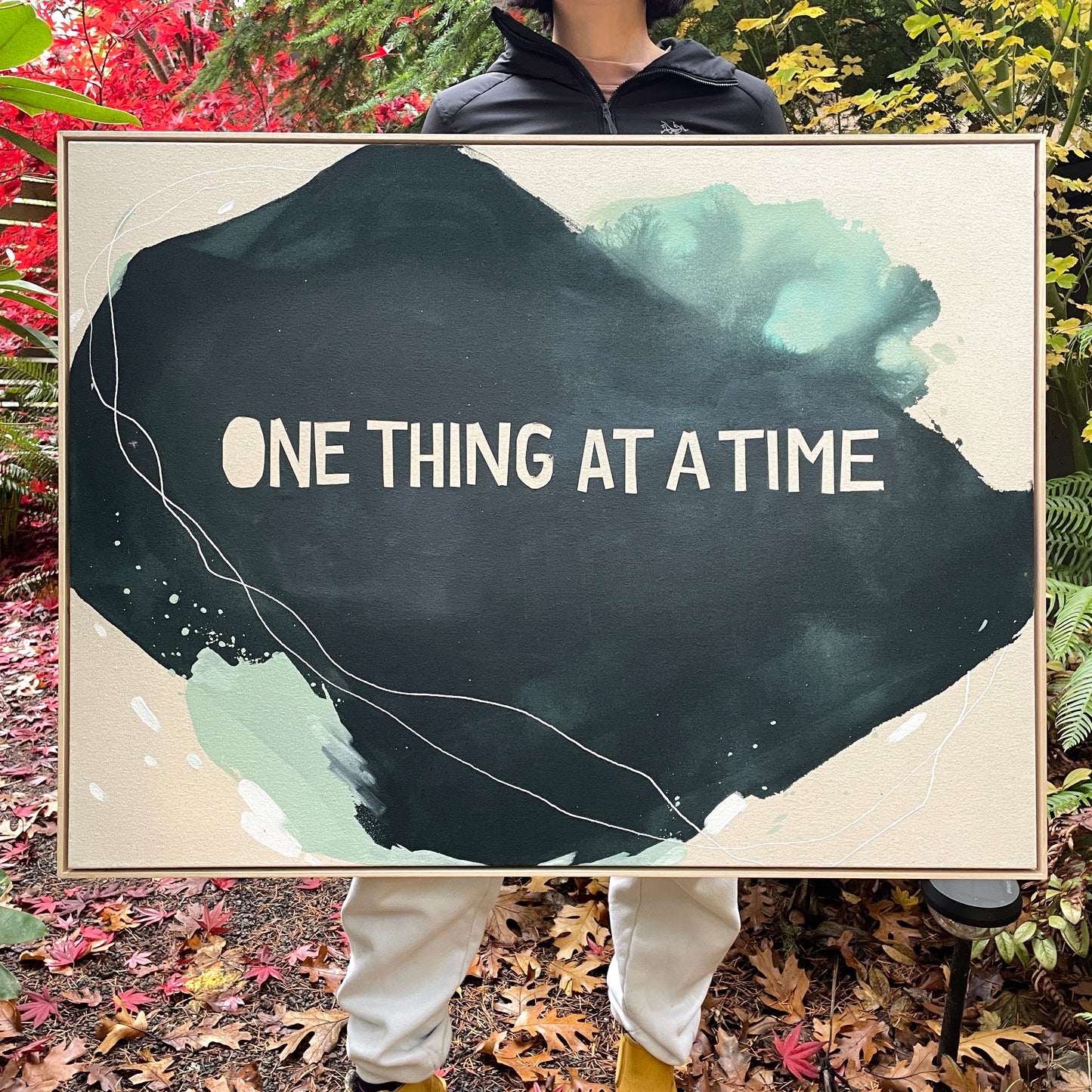 person holding close up photo of original abstract painting by britchida showing the framed canvas with navy, teal, and white shapes and white lines and the white text "ONE THING AT A TIME"