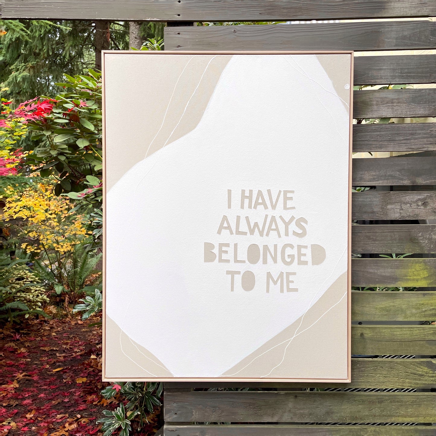 Large, framed, abstract painting with white shape in the center and encompassing lines with a tan background. In lower right of painting, the block text in tan reads: "I HAVE ALWAYS BELONGED TO ME"