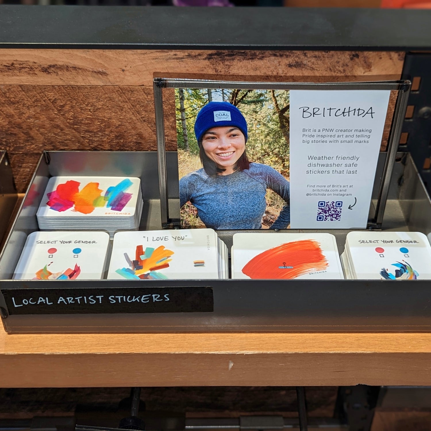 five piles of stickers by artist britchida in a metal box with the label "LOCAL ARTIST STICKERS" on a shelf at the REI flagship store in Seattle, Washington. A photo of britchida outdoors with a blue beanie hat and a description of the artist accompanies the stickers