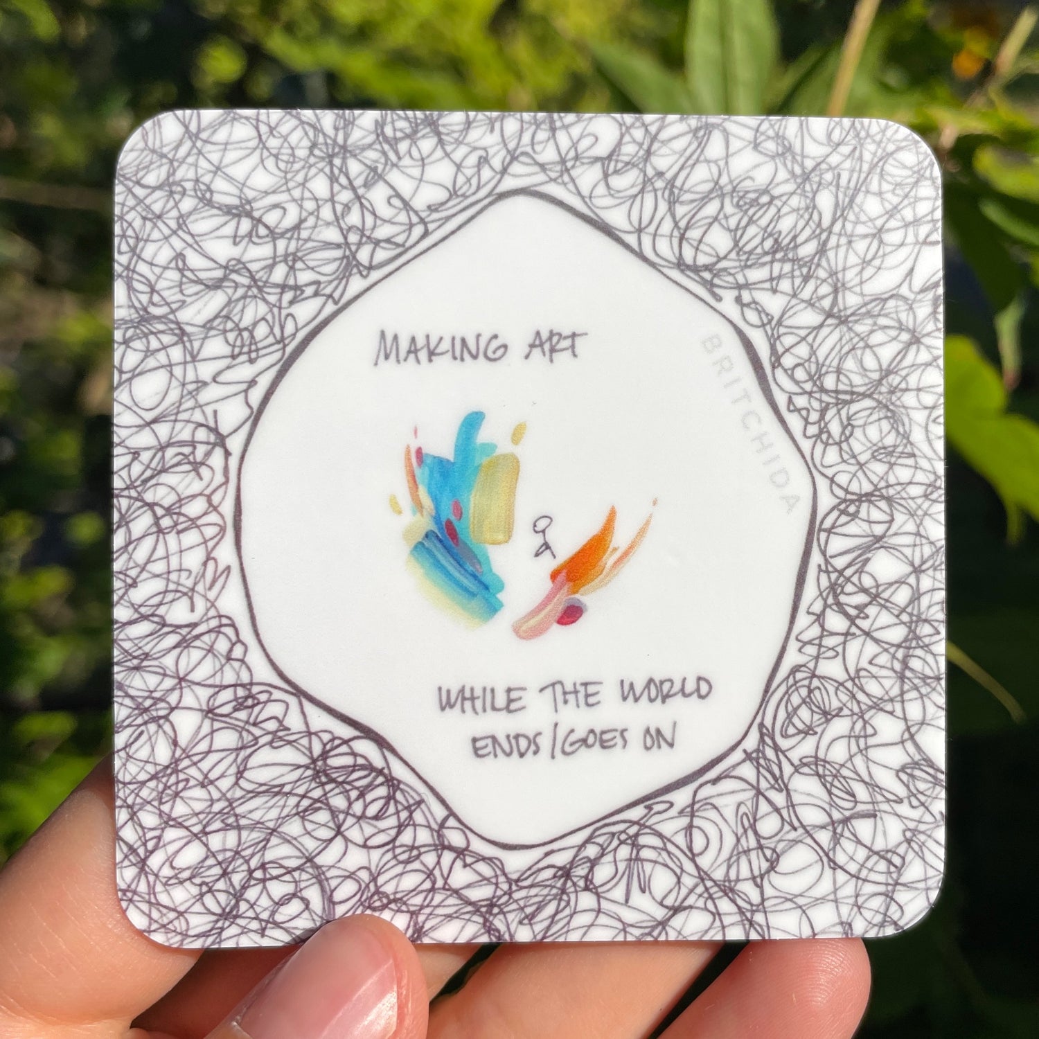 fingers holding small sticker titled "making art" by britchida with looping continuous black lines at the perimeter with a small stick figure surrounded by colorful paint strokes in the center. the text "MAKING ART WHILE THE WORLD ENDS/GOES ON" is inside the center area