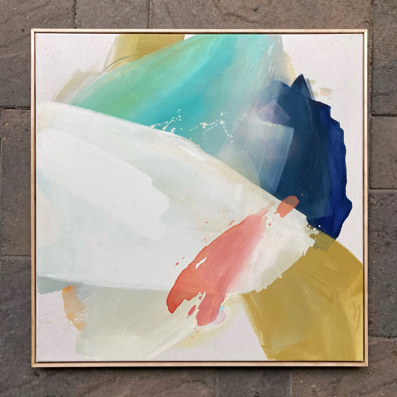 large framed abstract painting by britchida with blue, teal, yellow, pink, and white shapes. painting is laying on stone background