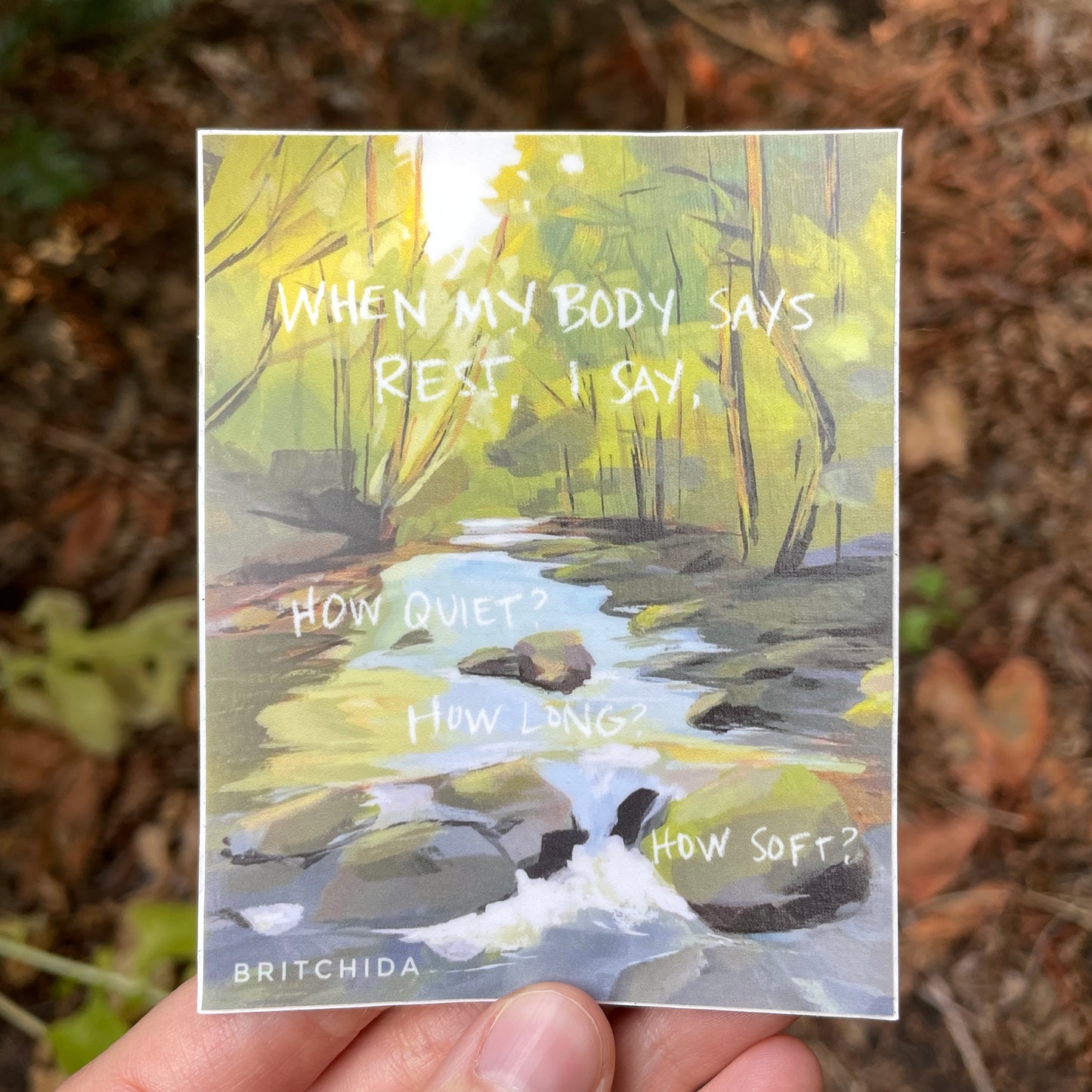 sticker of How Soft? print. painting of a creek in the forest with the creek and stones in the center bottom and green trees with brown trunks on the sides of the creek. The text "When my body says rest, I say, How quiet? How long? How soft?" in white text over the landscape