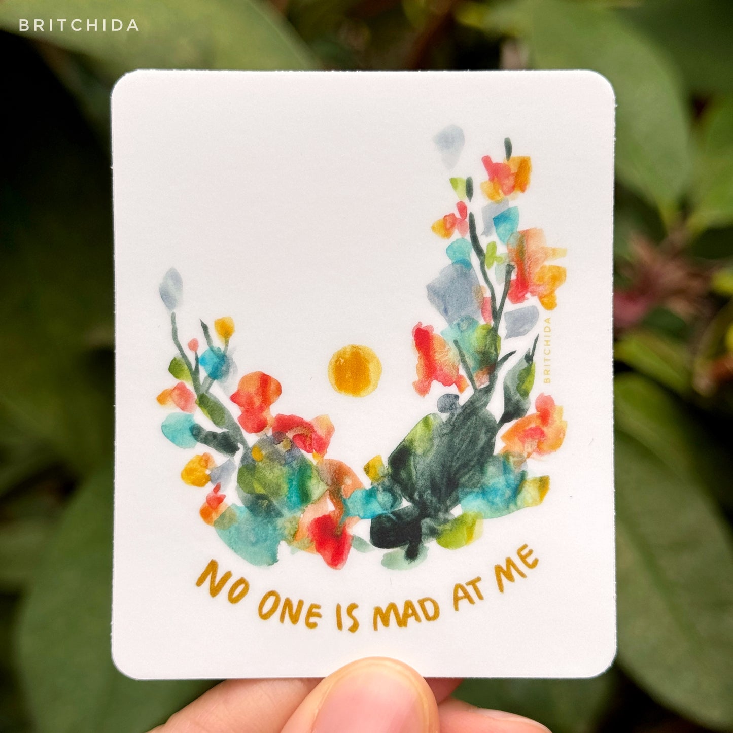 sticker of No One is Mad at Me print with abstract floral shapes in an arch with a single yellow dot in the middle of the upside down arch. At the bottom there is text that reads "no one is mad at me"