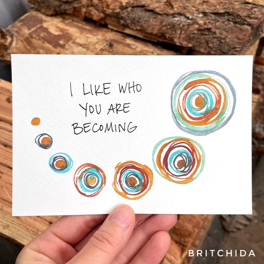 the text “I like who you are becoming” or "I like who I am becoming" is set above a swooping line of round shapes. Each shape has one ring larger than the one before, painted in a wet paint so the layers begin to blend a bit.