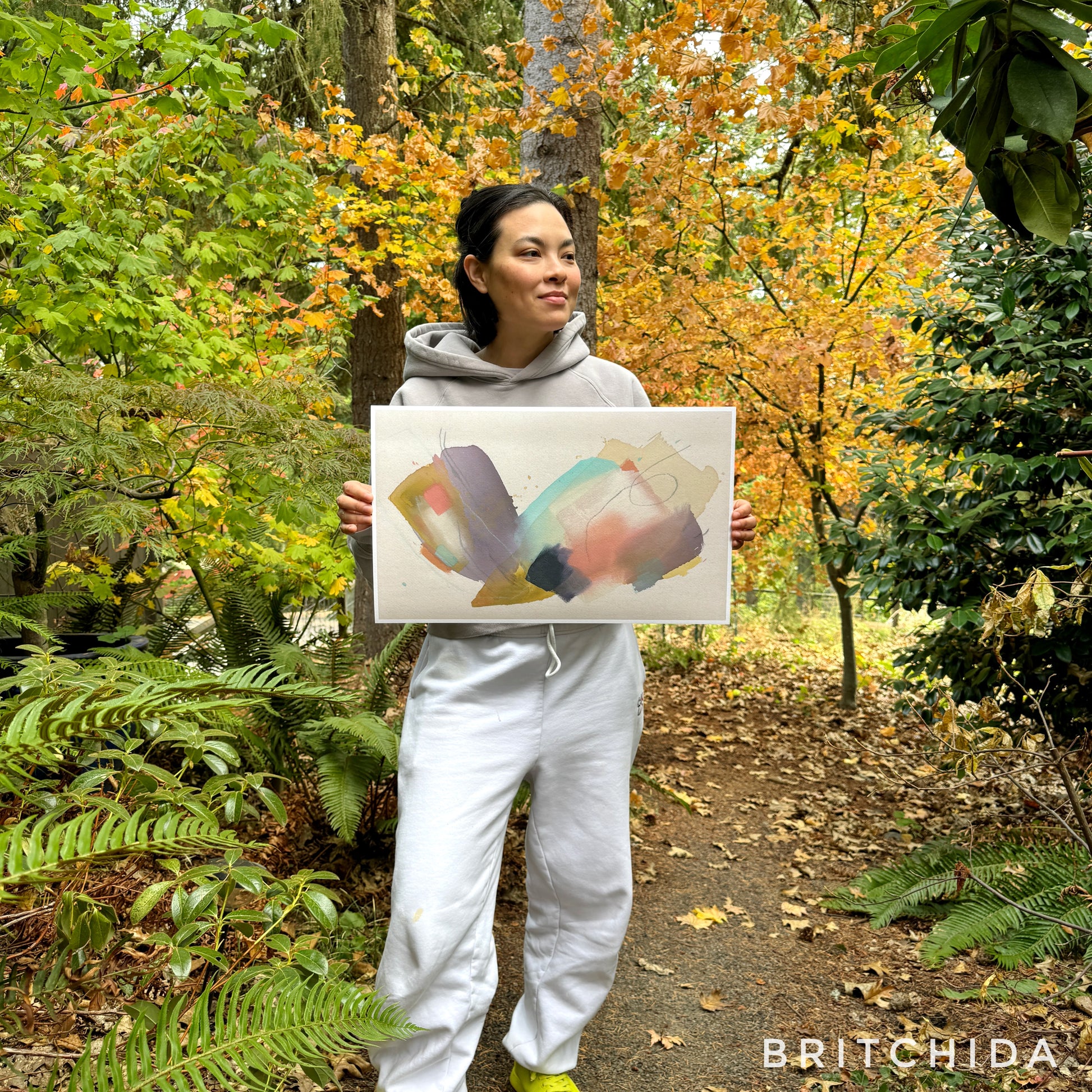 person with dark hair and ivory sweatsuit standing in fall foliage holding print of abstract painting by britchida with yellow, teal, peach, navy, and lilac shapes with various thin lines