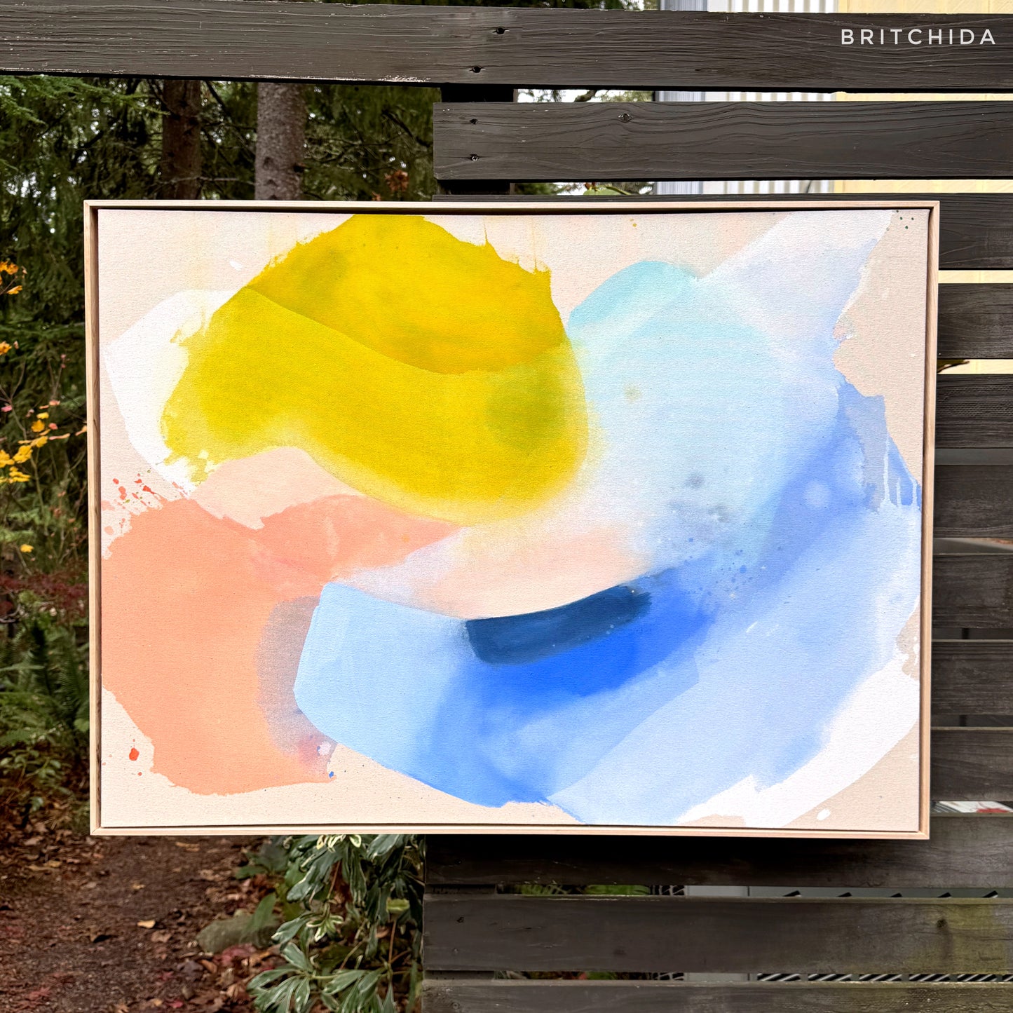 abstract painting by Britchida hanging on a wooden fence with various shapes of yellow, white, ice blue, blue, and pale orange