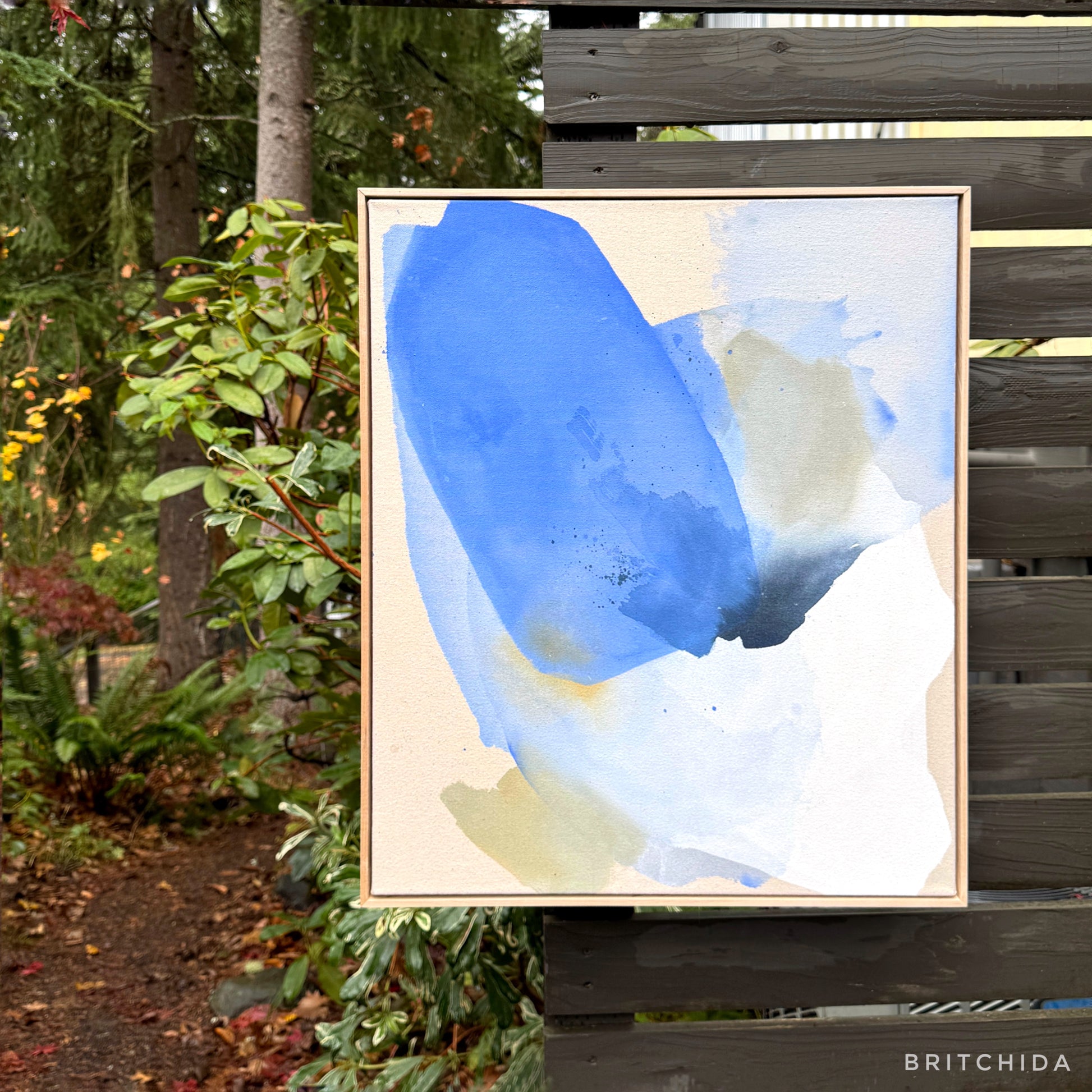 abstract painting by Britchida hanging on a wooden fence with various shapes of blue, gray, and white