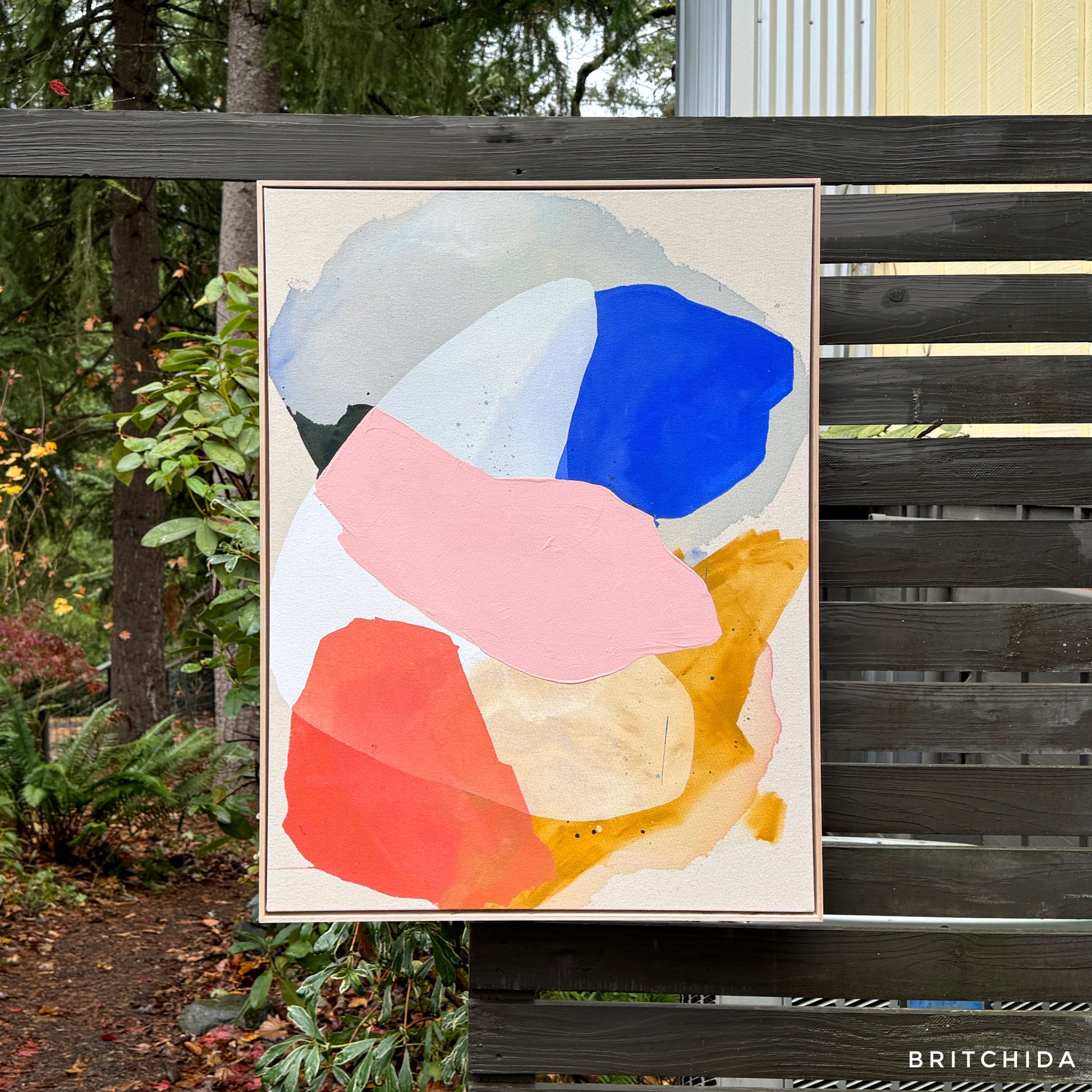 abstract painting by Britchida hanging on a wooden fence with various shapes of pink, white, gray, orange, red, and blue