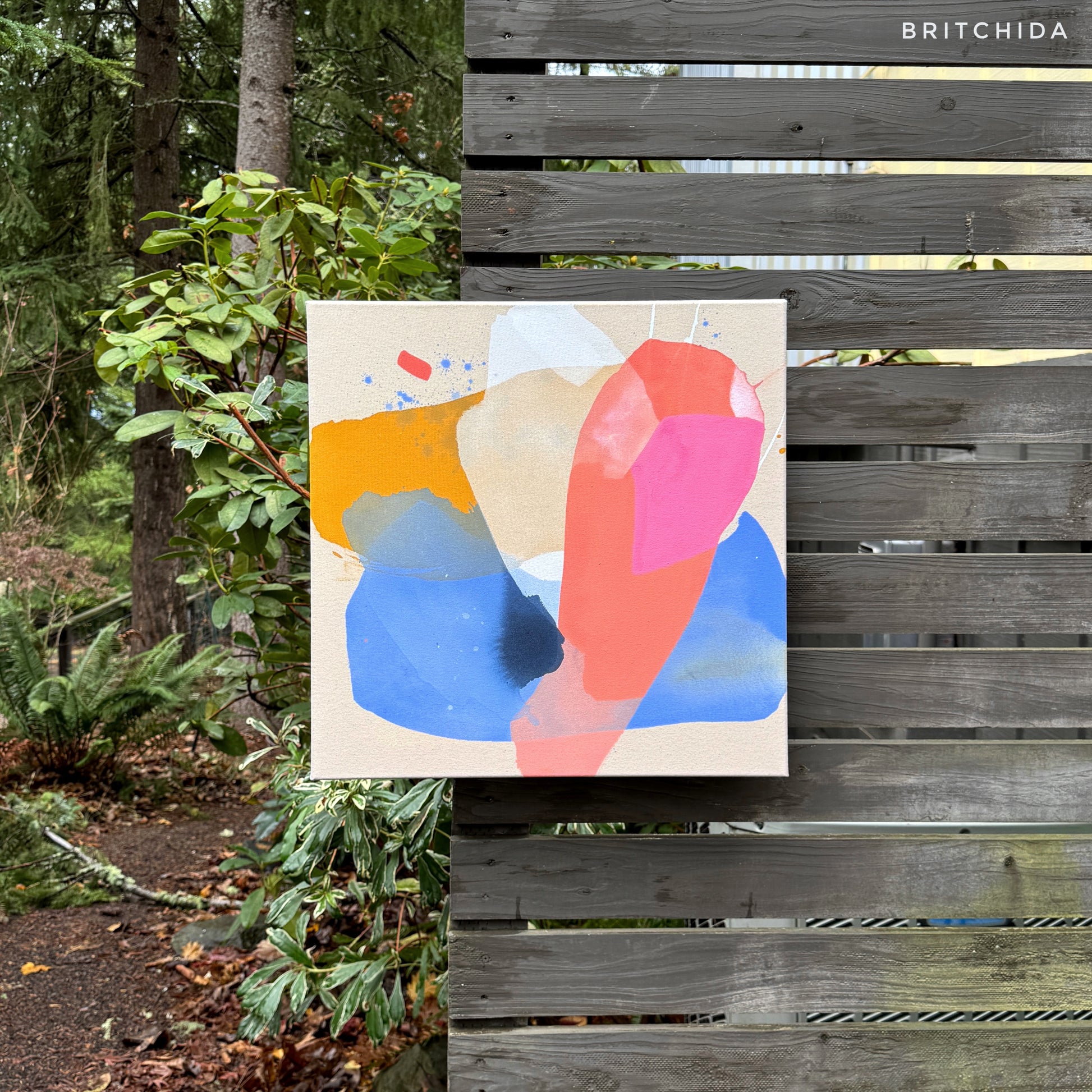 abstract painting by Britchida hanging on a wooden fence with various shapes of pink, blue, yellow, and white