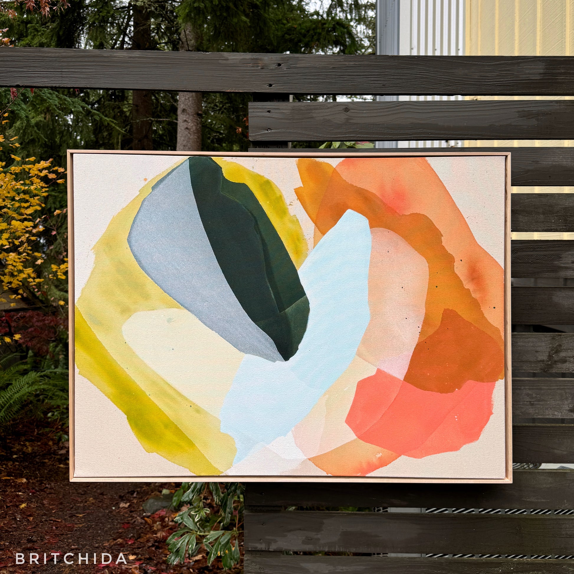 abstract painting by Britchida hanging on a wooden fence with various shapes of yellow, orange, pink, olive green, white, and ice blue