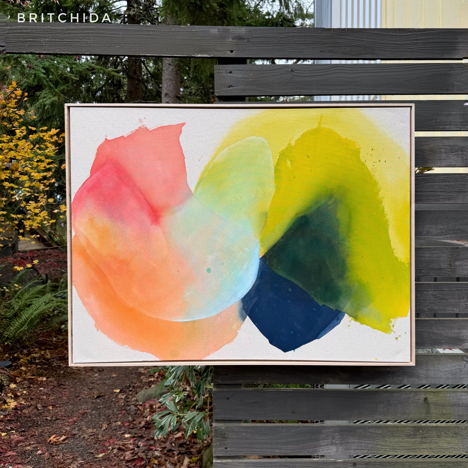 abstract painting by Britchida hanging on a wooden fence with various shapes of red, orange, ice blue, navy blue, and yellow