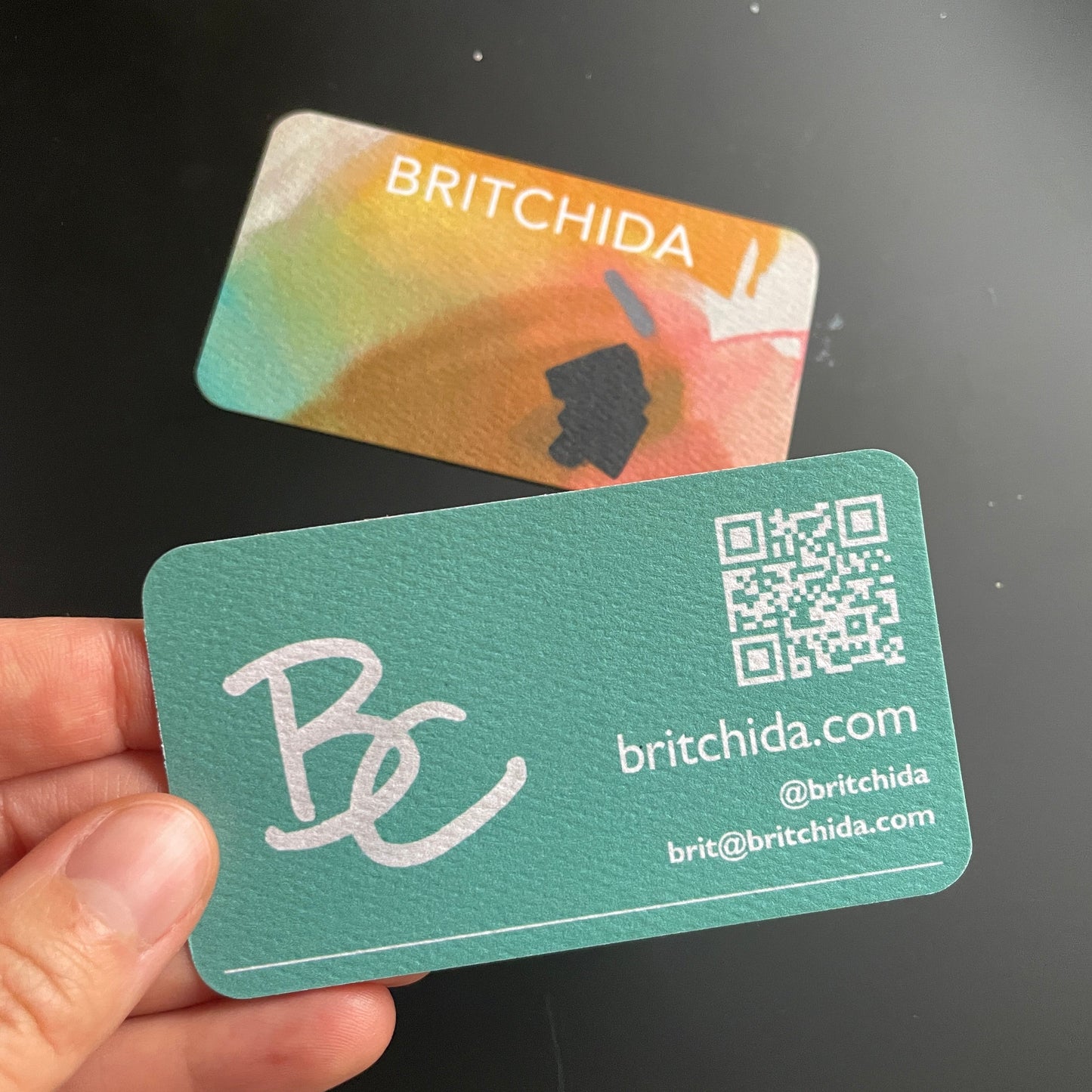 britchida business cards showing the front and back. the front (laying on the table) shows an abstract painting with orange, teal, black, gray, and pink shapes. the back is a teal color with the britchida logo (BC) and contact information