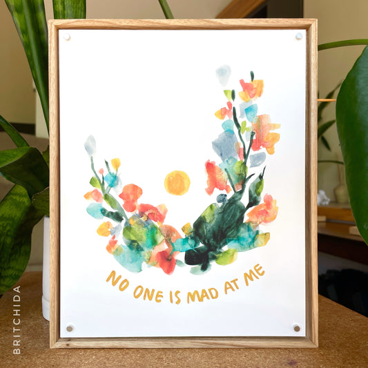 print "NO ONE IS MAD AT ME" in wooden frame with 4 small silver magnets holding print in frame. plants can be seen in the background