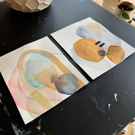 two abstract paintings by britchida with various shapes of pink, blue, tan, navy, and orange colors. paintings are laying on black desk 