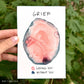 a large area of pink paint has a rim of dark blue around the edge which is blending a bit and bleeding in. At the top is the word “grief” and below the pink is labeled “loving you” and the blue “without you”