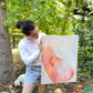 photo of person with dark hair, white long-sleeved shirt, and jean shorts standing in a garden holding original abstract painting by britchida with pink and white shapes and lines on canvas. a small white dog can be seen in the background sniffing the tree