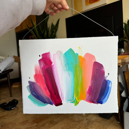 abstract pride flag with blue, green, orange, pink, red, orange, teal, and magenta colors on hanging mount. television and plants are visible in the background