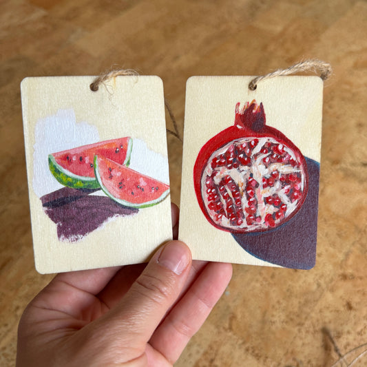 two wooden rectangular ornaments. the right ornament shows 2 slices of watermelons and the left ornament shows a cross-section of a pomegranate 