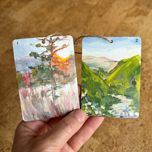 two wooden rectangular ornaments of landscape scenes. the left ornament shows a tree in a field and the right ornament shows a mountainous valley with a river winding between green mountains 