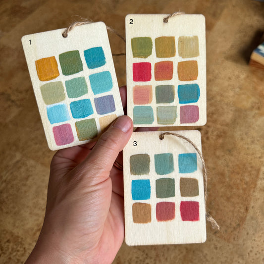 person holding 3 rectangular wooden ornaments with 12 squares of various colors on each resembling a paint palette