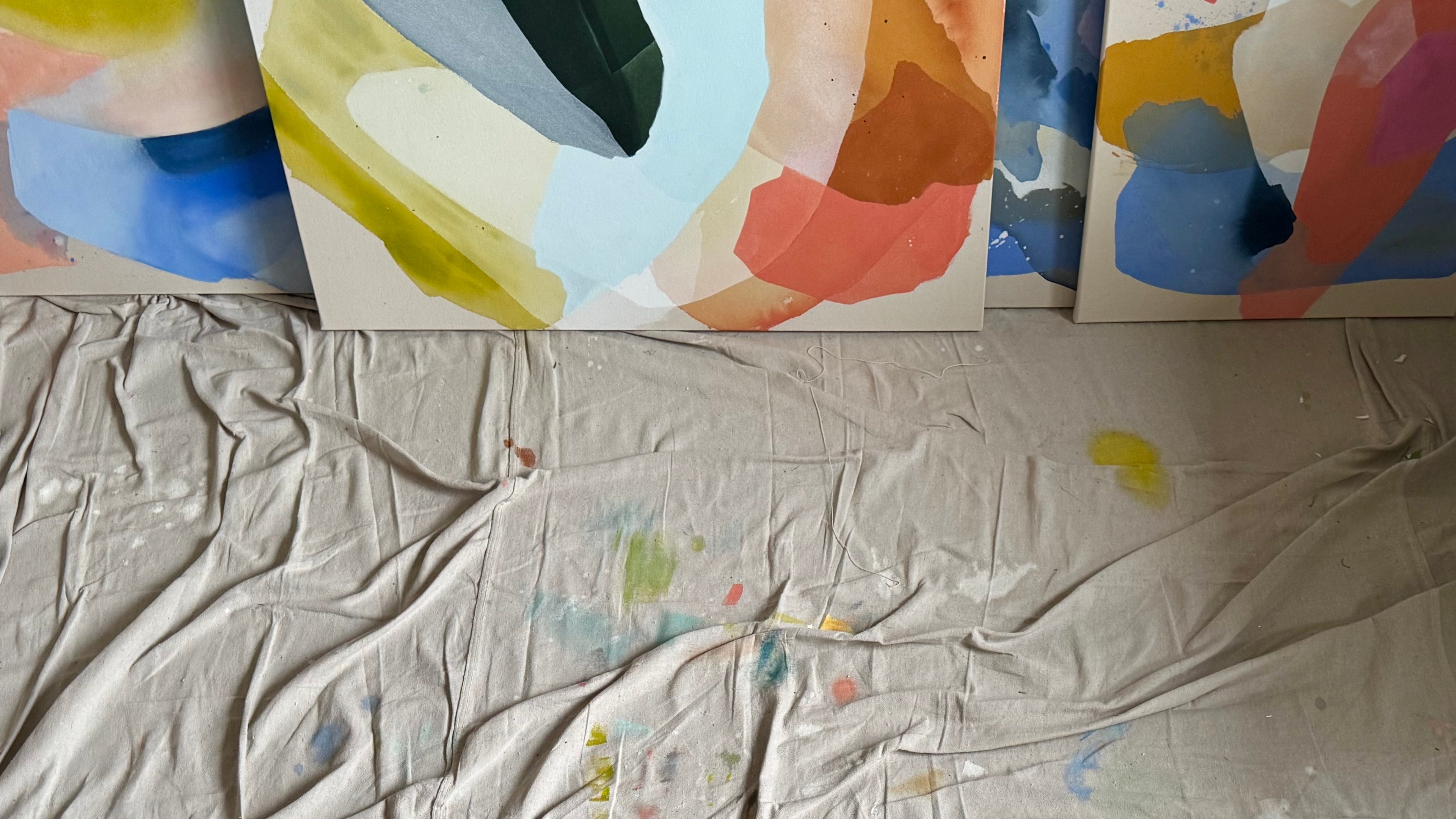 Paint splattered dropcloth with several colorful abstract paintings leaning on a wall