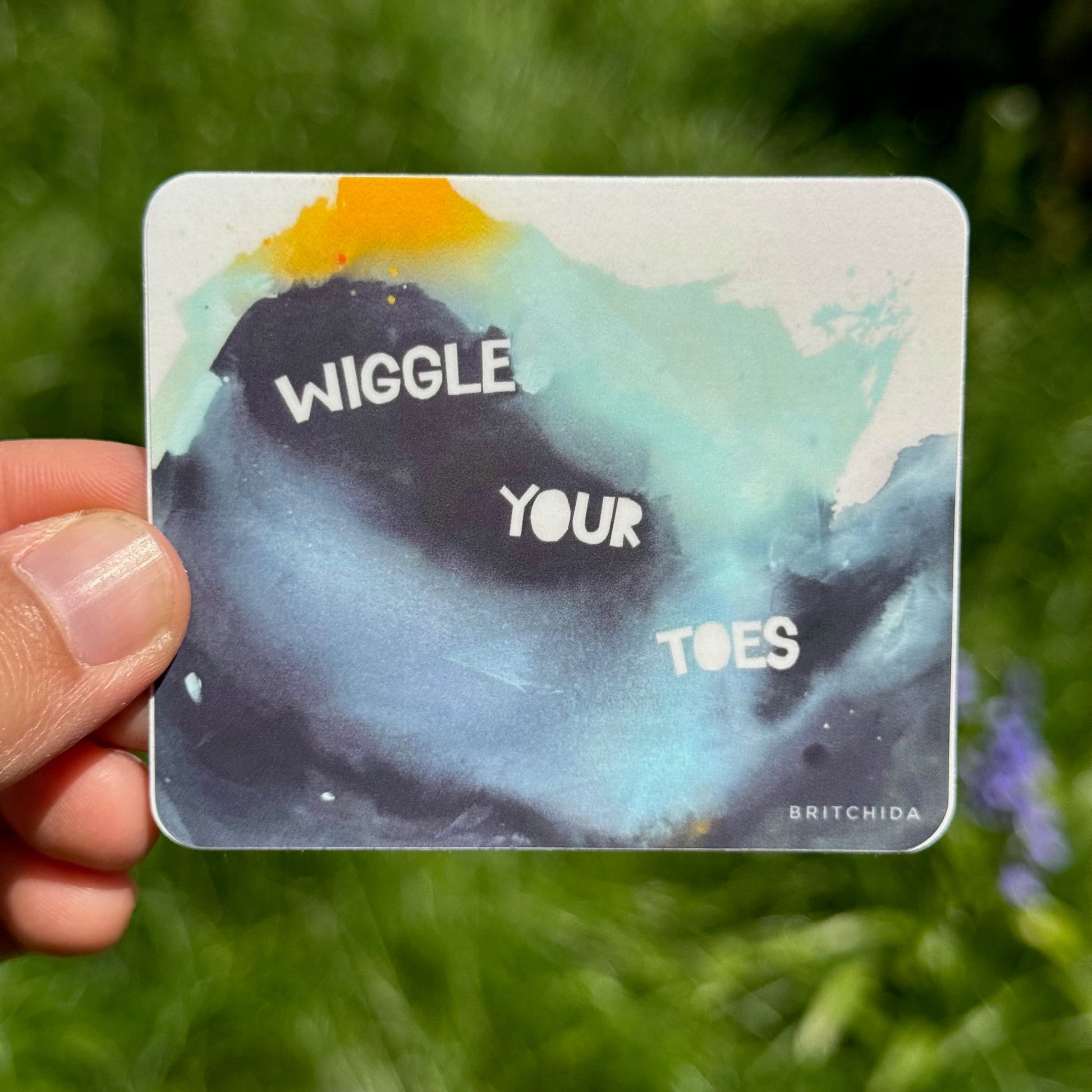 sticker of WIGGLE YOUR TOES original painting with navy, teal, and yellow watercolor paints in the background and white block letters spelling "WIGGLE YOUR TOES"