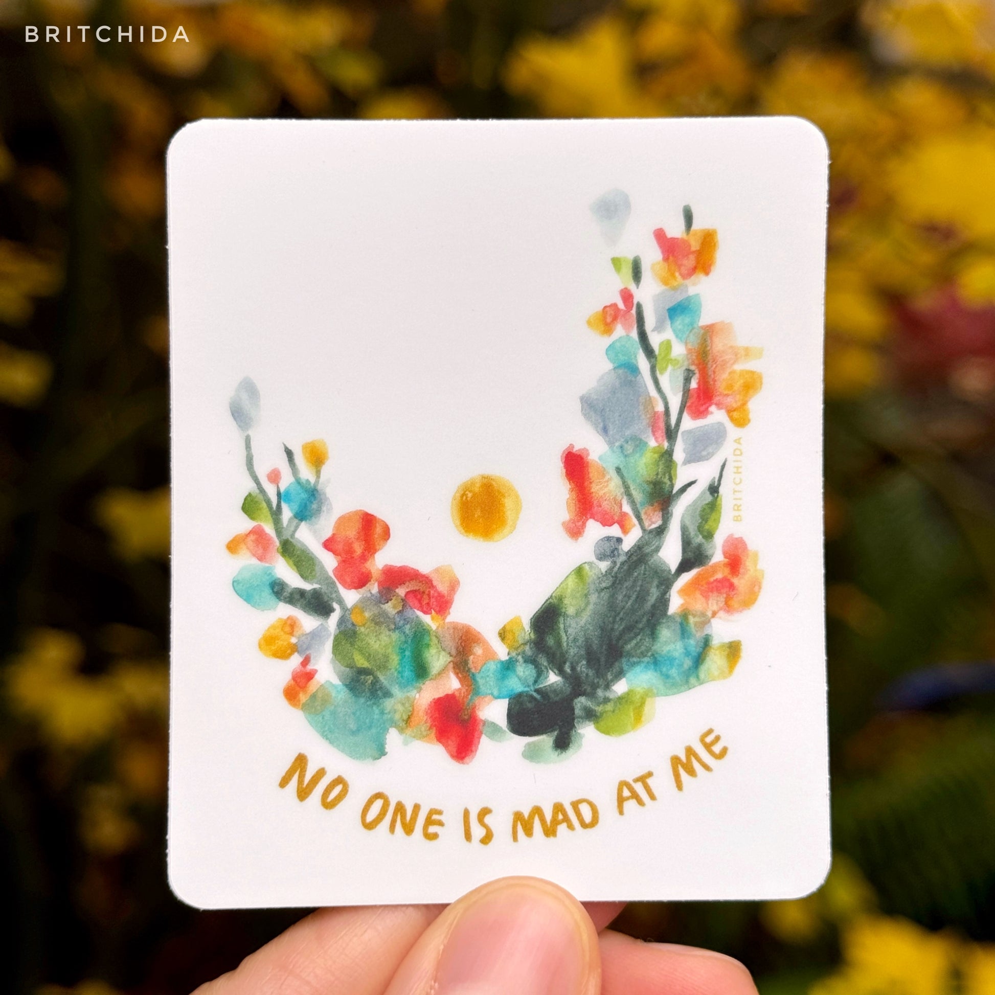 sticker of No One is Mad at Me print with abstract floral shapes in an arch with a single yellow dot in the middle of the upside down arch. At the bottom there is text that reads "no one is mad at me"