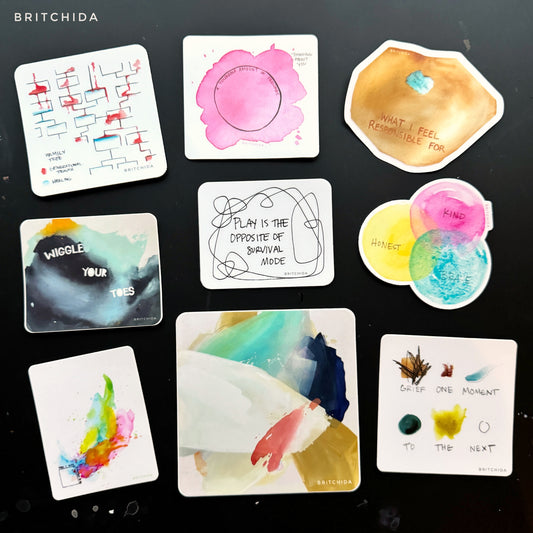 9 stickers (generational trauma, thinking about you, so responsible, wiggle your toes, play, kind honest brave, who I am, ease, and grief one moment to the next) on a black desk