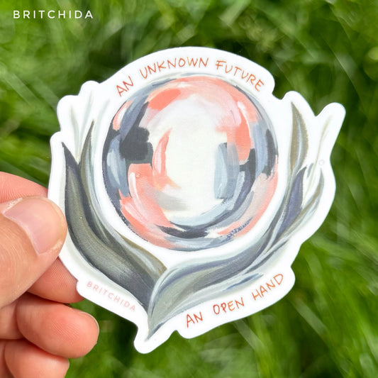 sticker of Unknown Future print with a blue, pink, and white circle in the center with wispy green gray petals at the base appearing to hold the circle. At the top reads "an unknown future" and at the bottom the text says "an open hand"