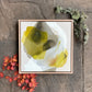 framed original abstract painting by britchida with yellow, white, and gray shapes with thin lines on tan canvas laying on stone floor with orange japanese maple leaves in lower left corner and mossy and lichen covered branch in upper right corner