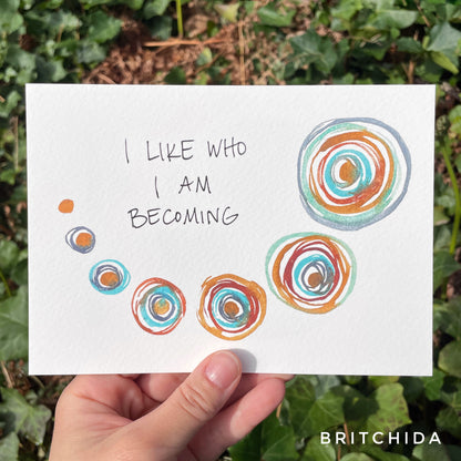 the text “I like who you are becoming” or "I like who I am becoming" is set above a swooping line of round shapes. Each shape has one ring larger than the one before, painted in a wet paint so the layers begin to blend a bit.