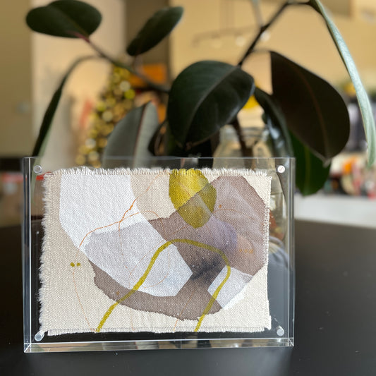 small abstract painting with gray, yellow, and white shapes on raw-edge canvas in clear acrylic frame with plants in the background