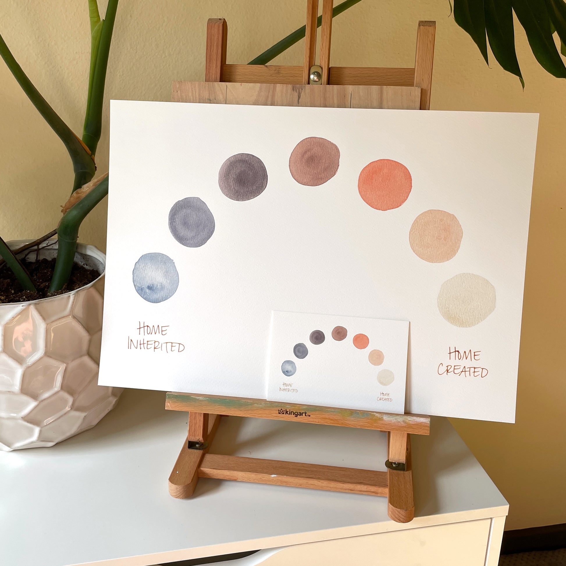 smaller print laid on top of larger print on wooden easel. print description: seven circles in a spectrum of soft neutral colors makes an arch with the words “home inherited” on the left and “home created” on the right.