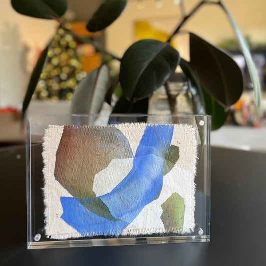 small abstract painting with blue, gray, and dark green shapes on raw-edge canvas in clear acrylic frame with plants in the background