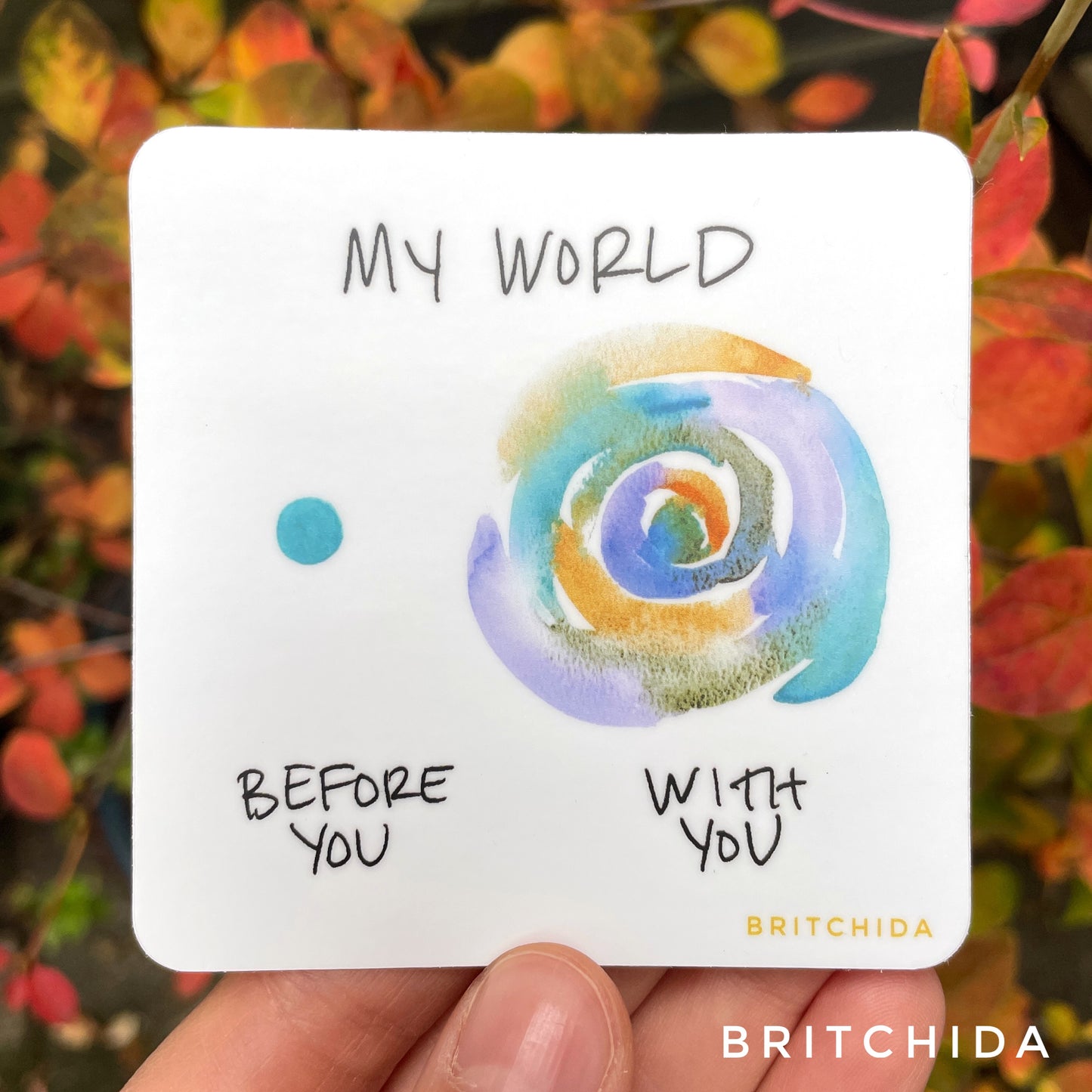 sticker of My World print with a single blue dot on the left with the text "before you" and a larger shape of concentric circles of blue, purple, and orange and the text "with you" below. At the top is the text "my world"